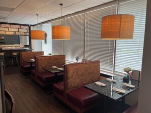 Um restaurante com assentos em cabines, janelas amplas com persianas e luminárias pendentes.