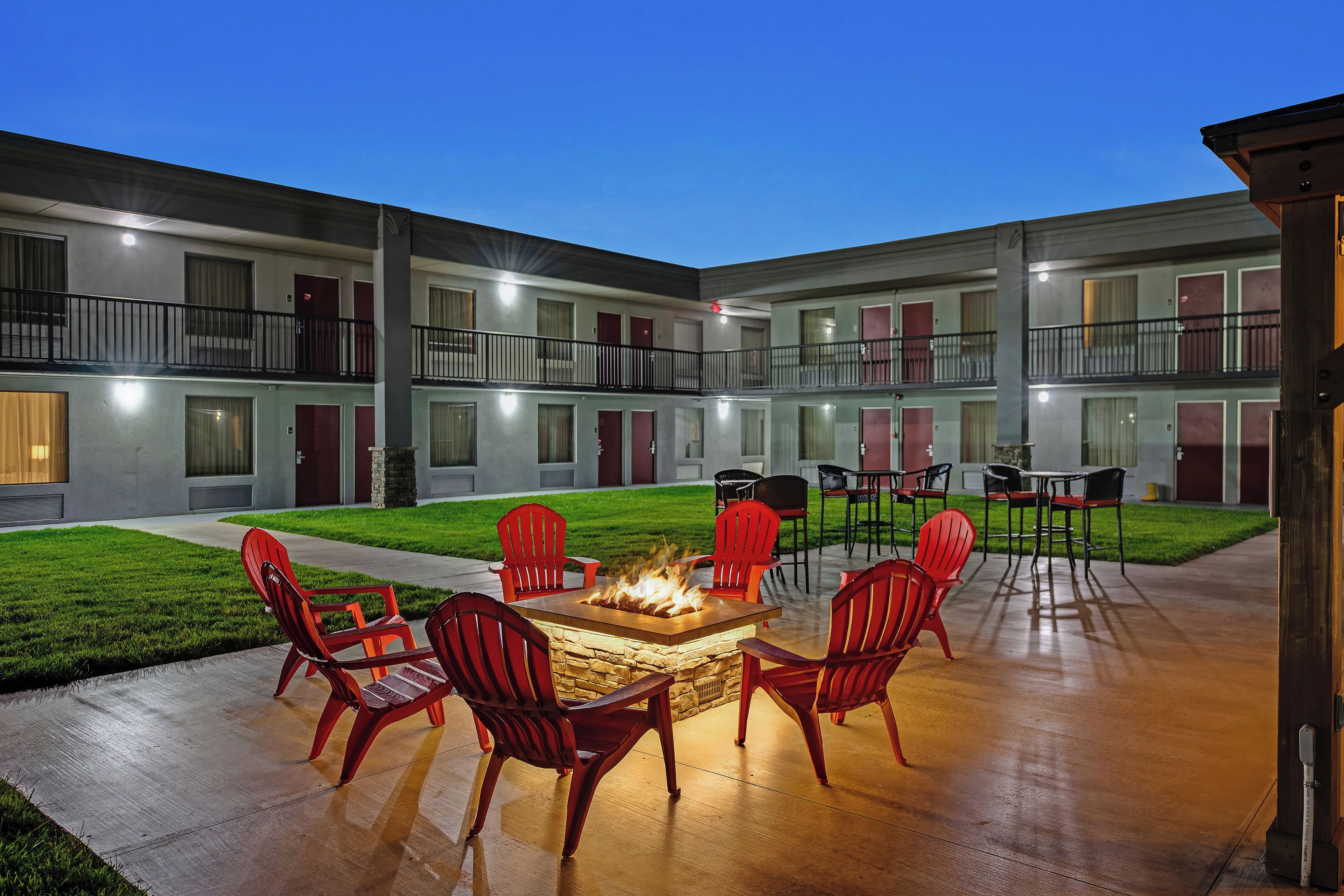 A courtyard with a fire pit surrounded by red chairs and tables, a grassy lawn, and a building with multiple balconies and doors.