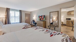 A hotel room with two beds, a desk, a chair, a TV, and a kitchen area visible through an open door.