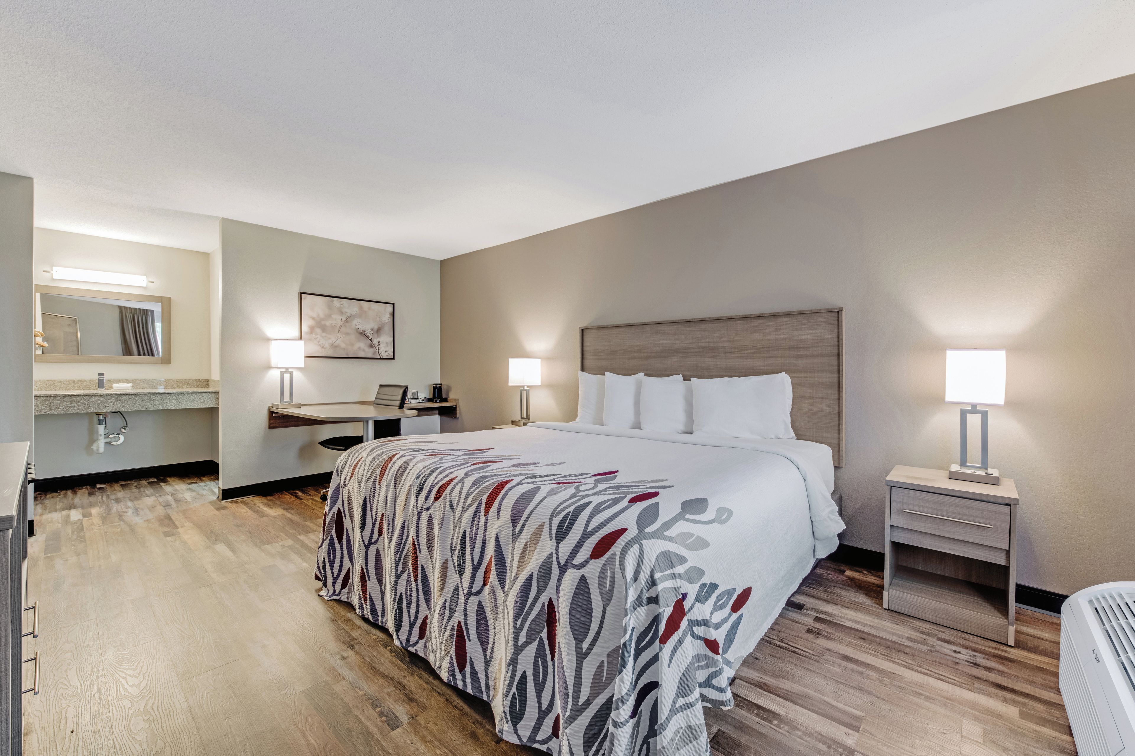 Habitación de hotel con cama, mesita de noche, lámpara, escritorio, silla y baño con espejo y lavabo.