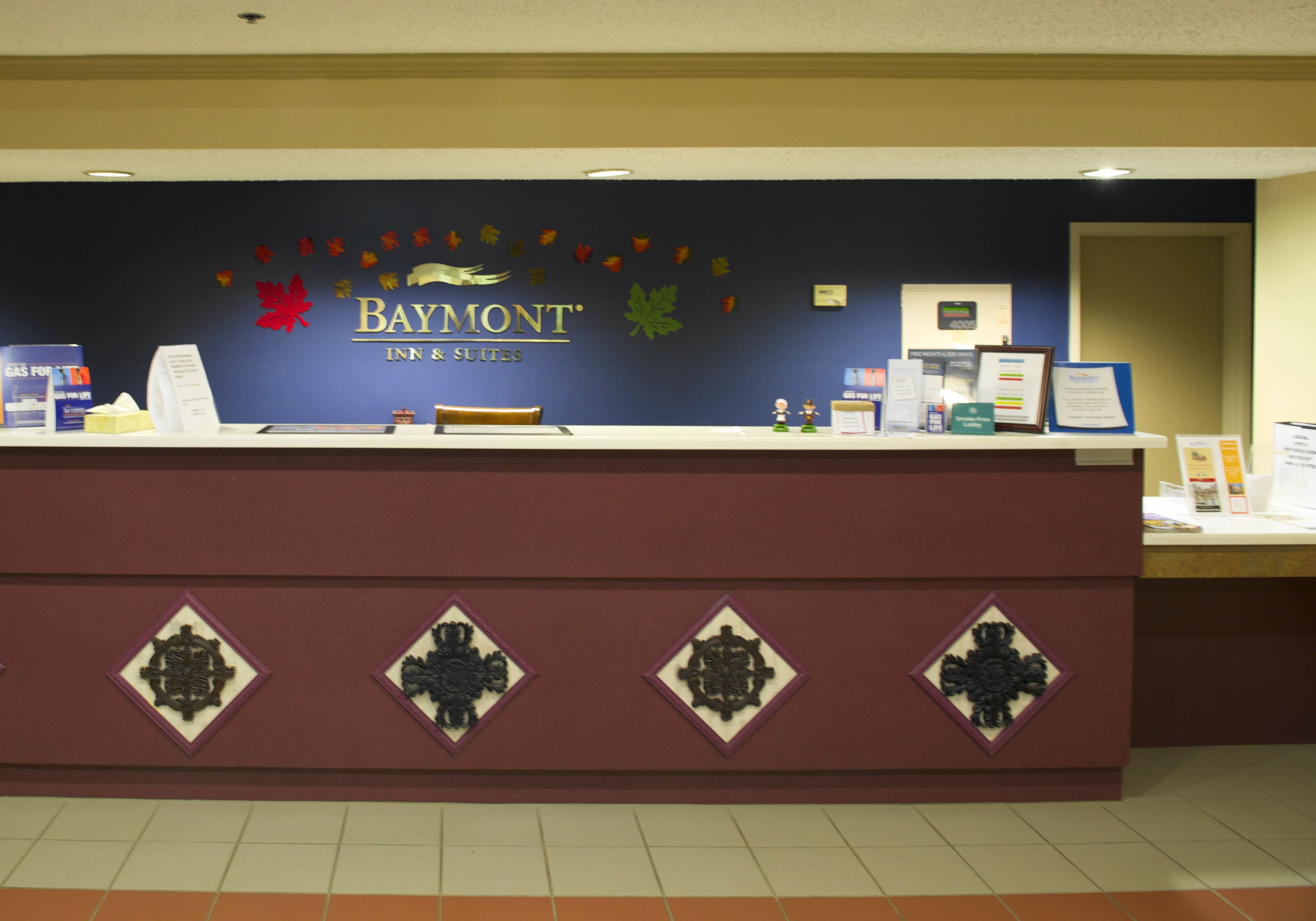 A reception desk at Baymont Inn & Suites with a dark blue wall, a sign with autumn leaves, and a row of framed certificates.