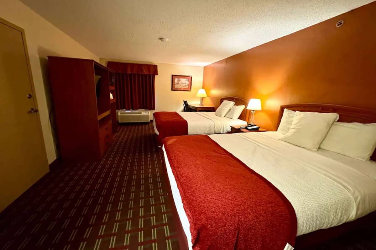 Une chambre d’hôtel avec deux lits, une moquette rouge, une armoire en bois, un bureau avec une lampe et un climatiseur fixé au mur.