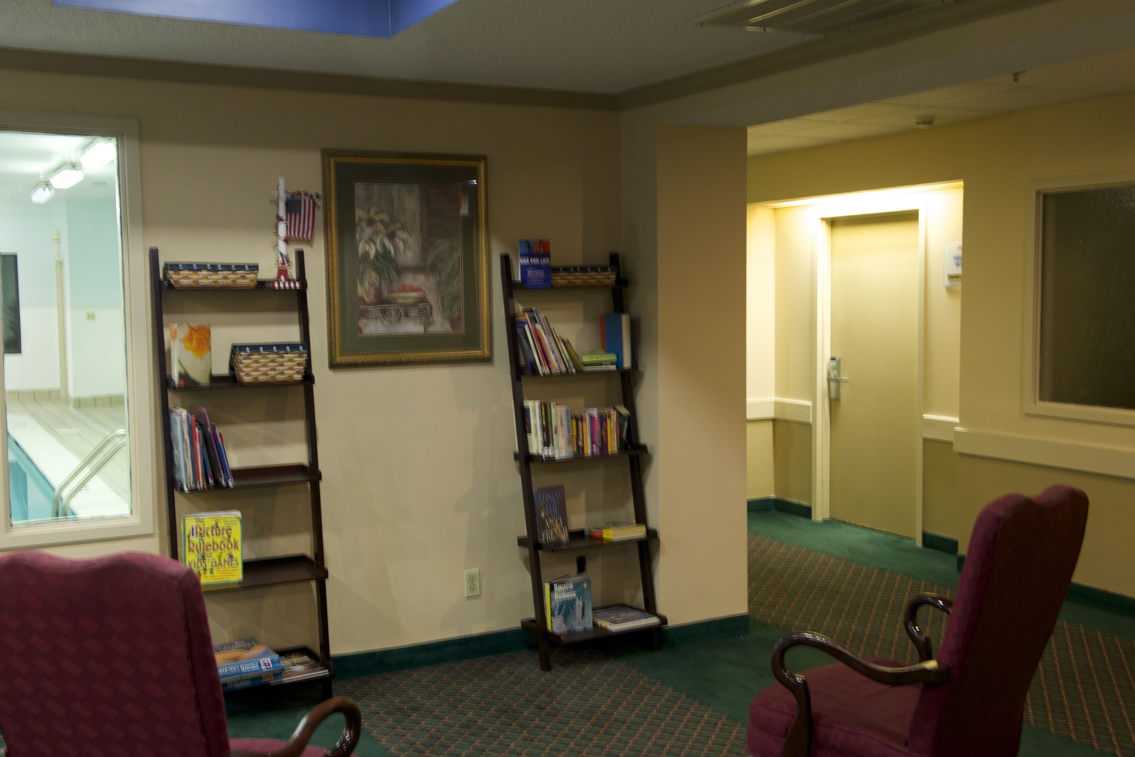 Um saguão de hotel com chão coberto de carpete, duas estantes de madeira com livros, um quadro emoldurado e duas cadeiras vermelhas.