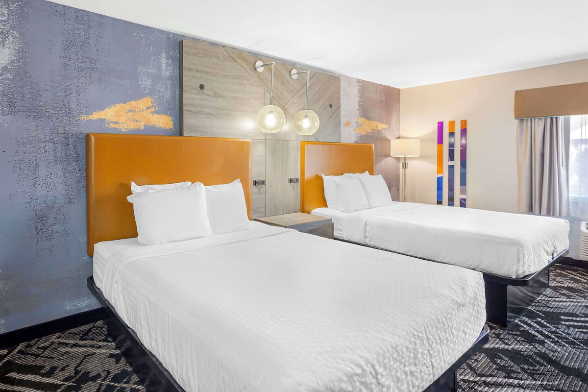 A hotel room with two beds, a large mirror, a modern light fixture, and a view of the city.