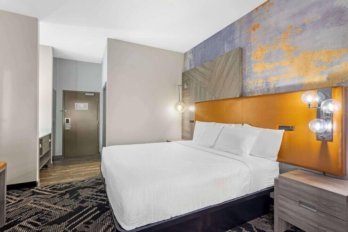 A hotel room with a bed, bedside table, wall-mounted light, and a door leading to a hallway.