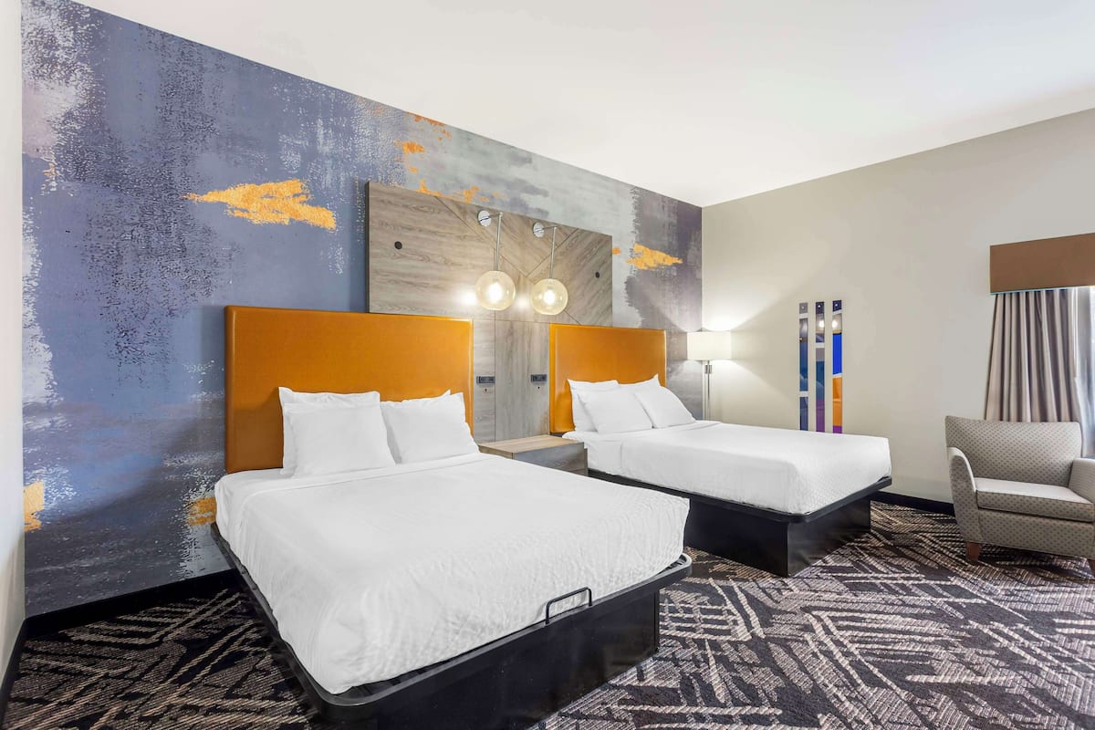 A hotel room with two beds, a patterned carpet, a modern wall with a textured design, and a chair.