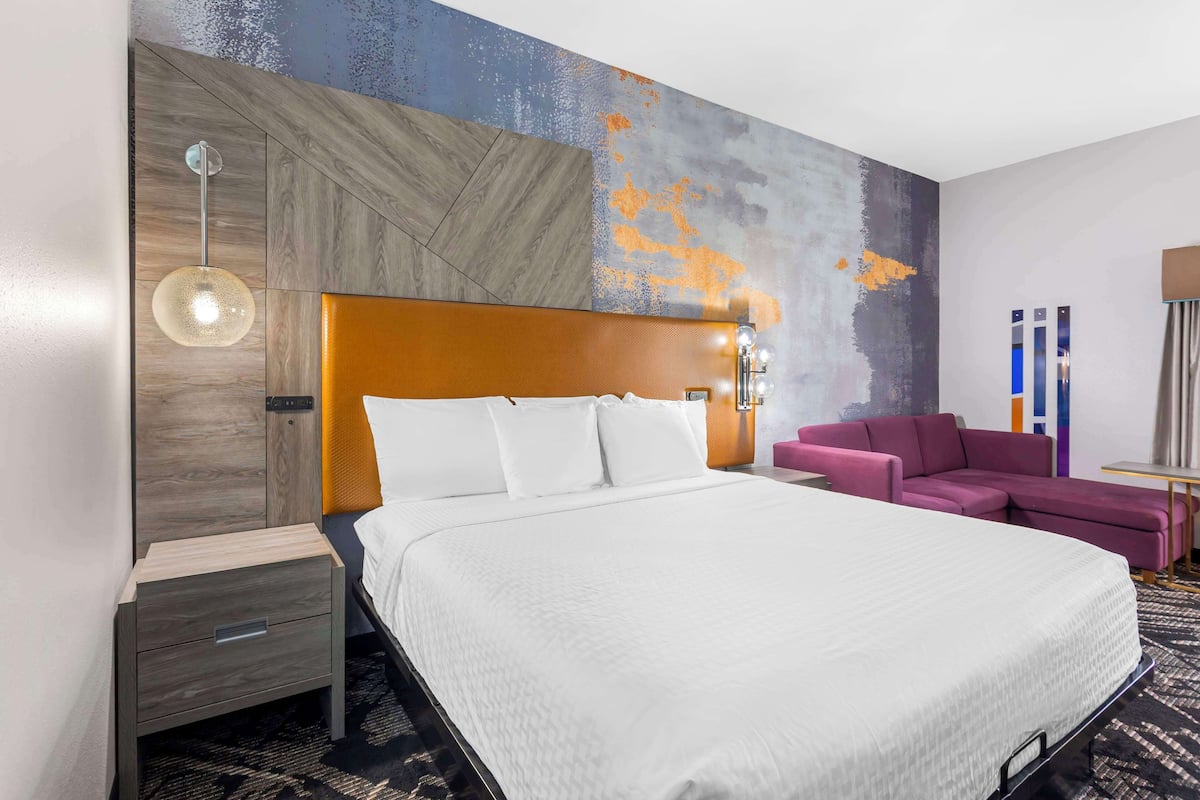 A modern hotel room with a large bed, a wooden headboard, a purple sofa, and a wall with a textured mural.