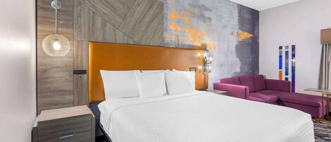 A modern hotel room with a large bed, a wooden headboard, a purple sofa, and a wall with a textured mural.