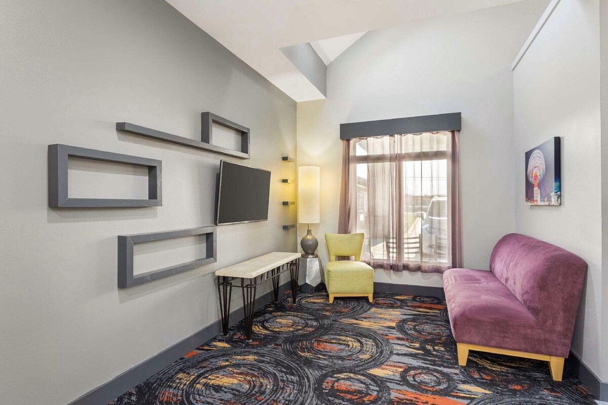 A hotel room with a TV, a small table, a chair, a sofa, and a patterned carpet.