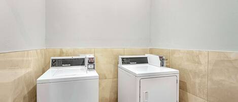 Una sala de lavandería con dos lavadoras blancas de carga frontal y una pared de azulejos color beige.