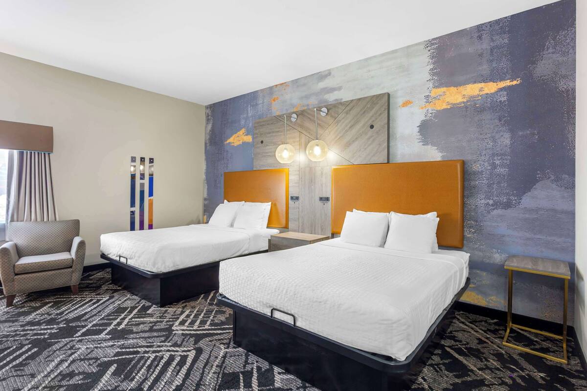 A hotel room with two beds, a chair, a small table, and a wall with a textured mural.