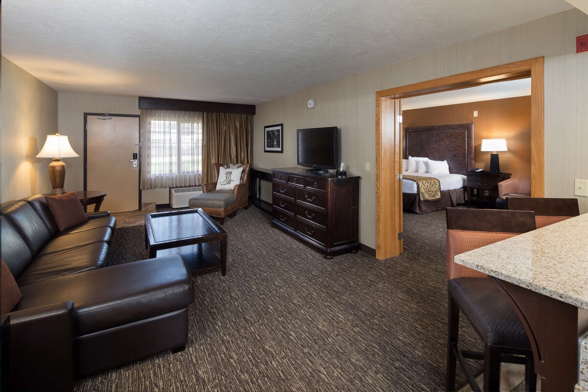 Habitación de hotel con sala de estar que incluye un sofá, una mesa de centro, un televisor, un aparador y un comedor con una mesa y sillas.