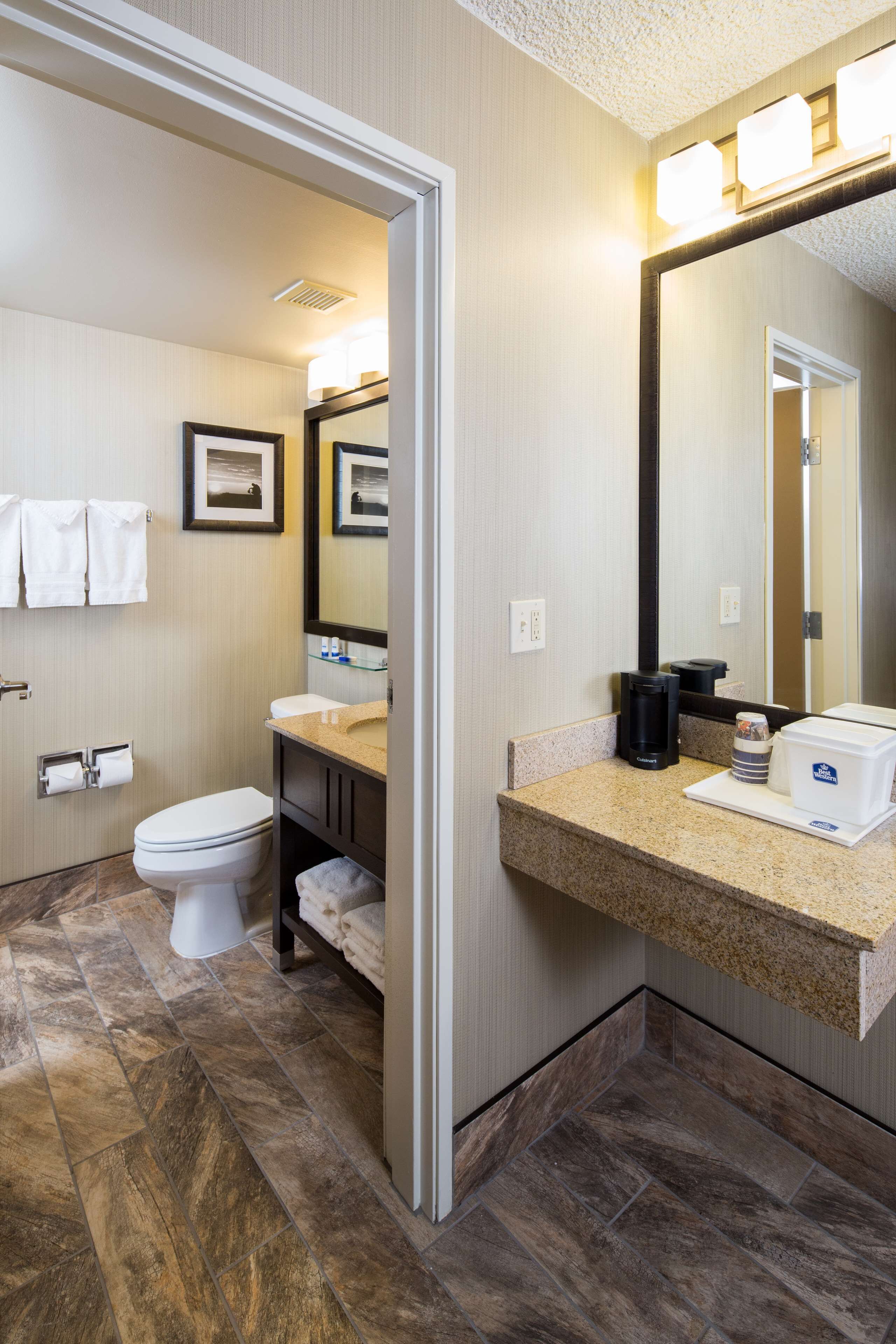 A bathroom with a toilet, sink, and mirror. There are towels hanging on the wall and a coffee maker on the counter.
