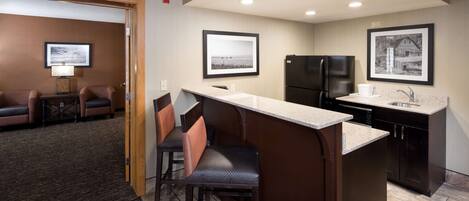 A compact kitchen with a bar area, a refrigerator, and a sink. There are framed pictures on the walls and a small seating area with chairs.