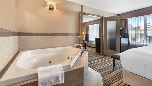 A hotel room with a large bathtub, a bed, a desk, and a view of the city through large windows. - Best Western Plus Calgary Centre Inn (Calgary)