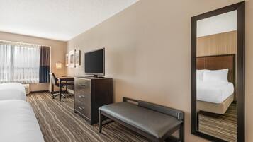 Habitación de hotel con cama, escritorio con silla, televisor, banco y un espejo grande.
