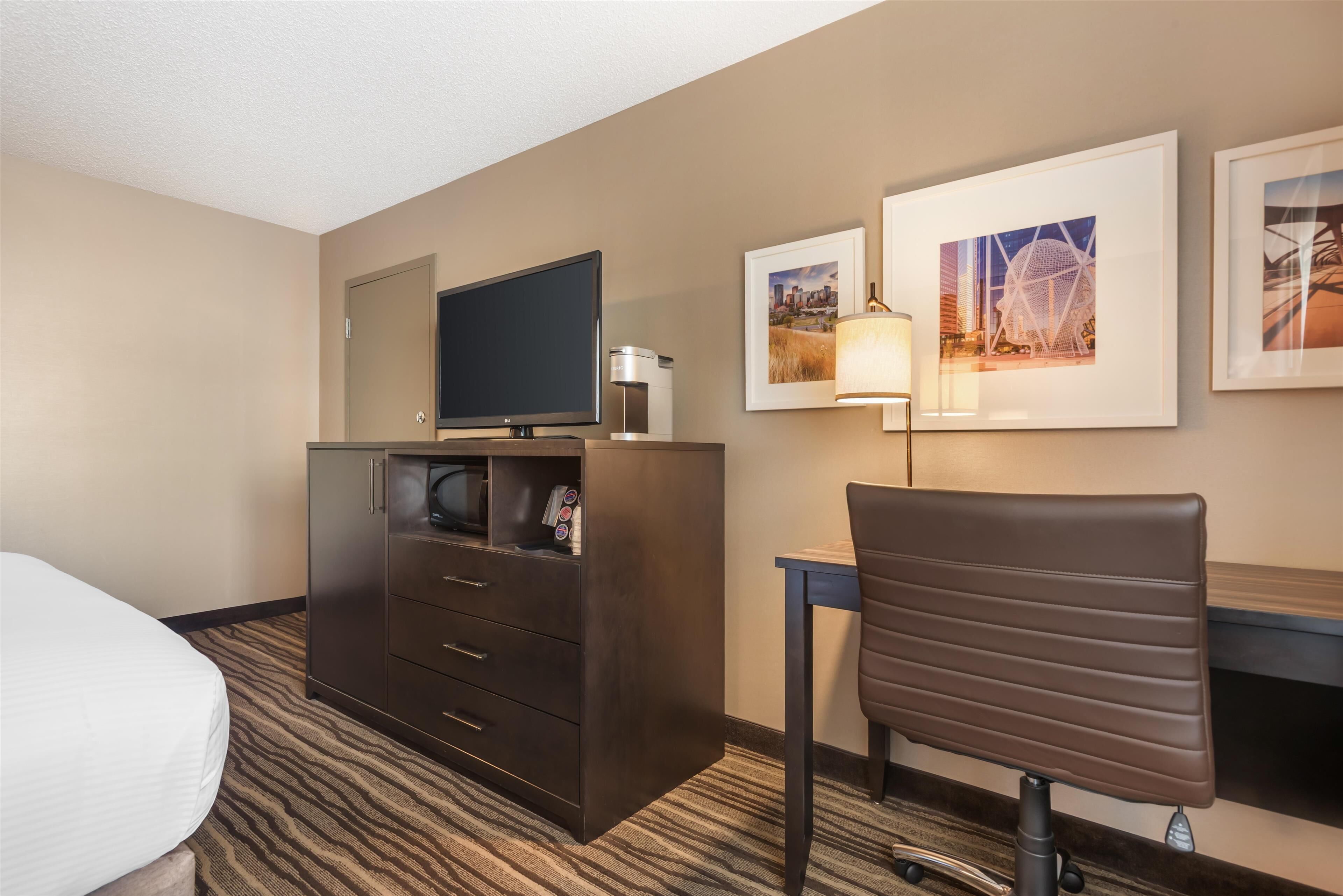 A hotel room with a bed, a desk with a chair, a television, a dresser, and framed pictures on the wall.