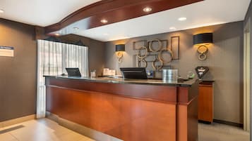 Un mostrador de recepción de diseño moderno, con acabado de madera y superficie de vidrio. Hay dos lámparas de pared con diseños circulares, y un letrero que indica el nombre del hotel y sus servicios.