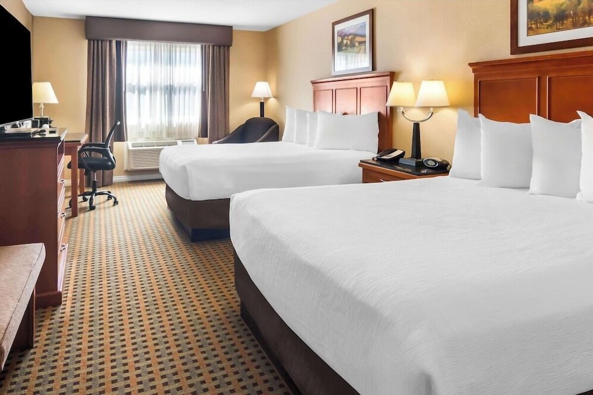A hotel room with two beds, a desk with a chair, a TV, a lamp, and a window with curtains.