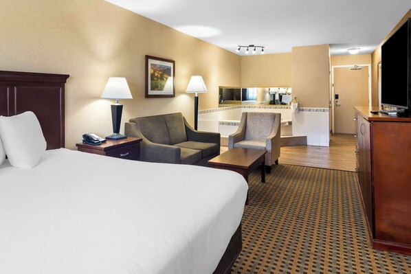 A hotel room with a large bed, two armchairs, a small table, a desk with a lamp, a telephone, and a television.
