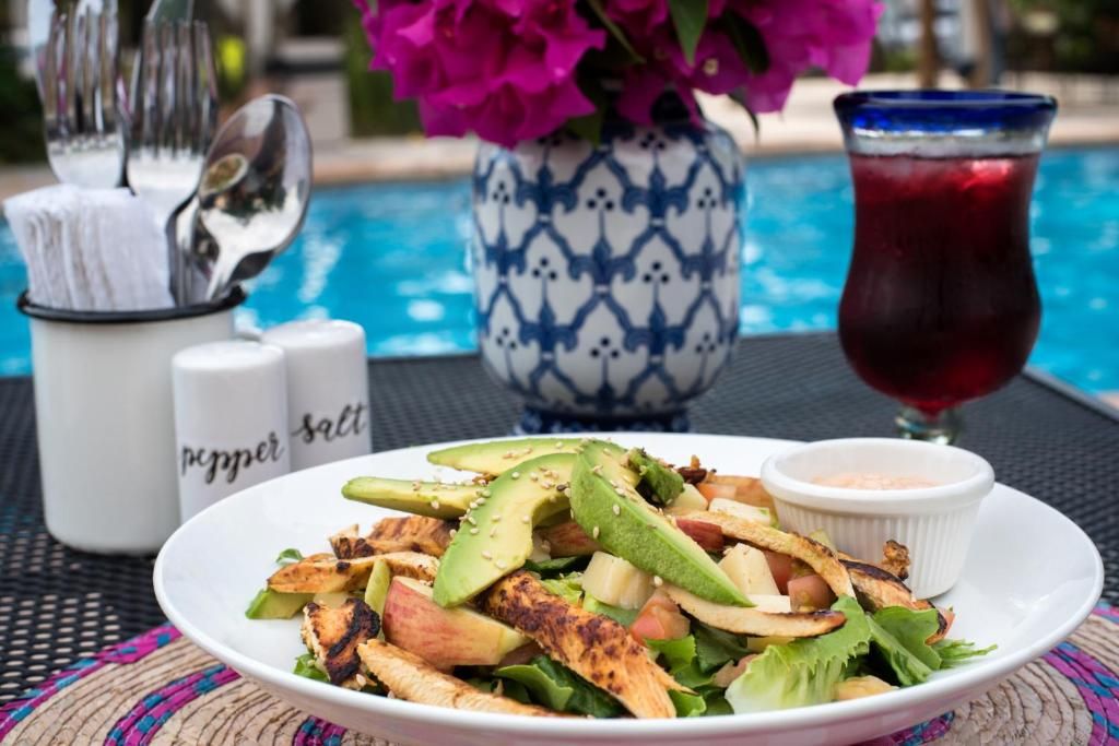 A plate of grilled chicken and vegetables with a side of sauce, a glass of red drink, and a vase of flowers.