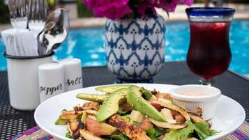 A plate of grilled chicken and vegetables with a side of sauce, a glass of red drink, and a vase of flowers.