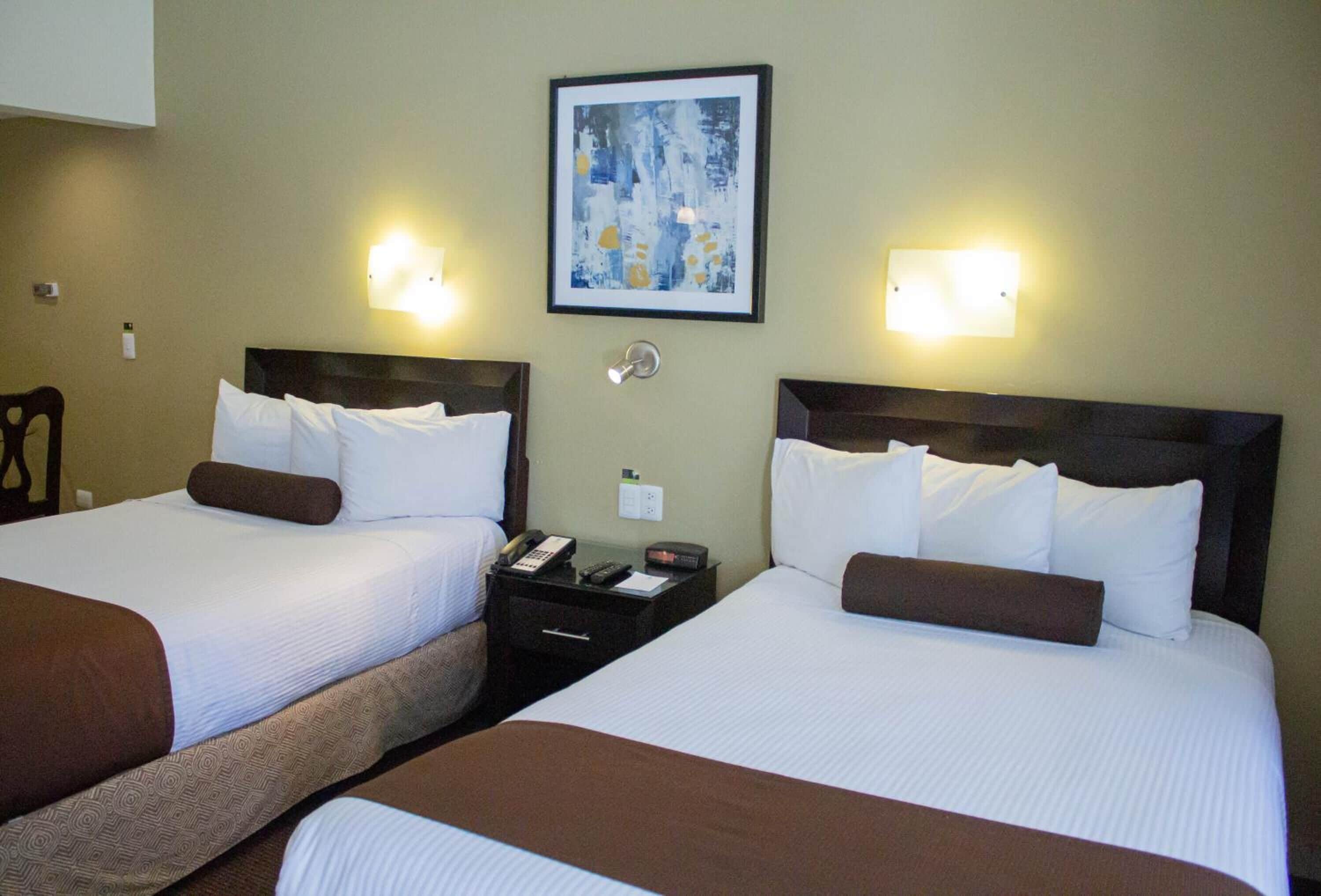 A hotel room with two beds, each with white linens and brown headboards. There is a framed abstract painting on the wall above the beds.