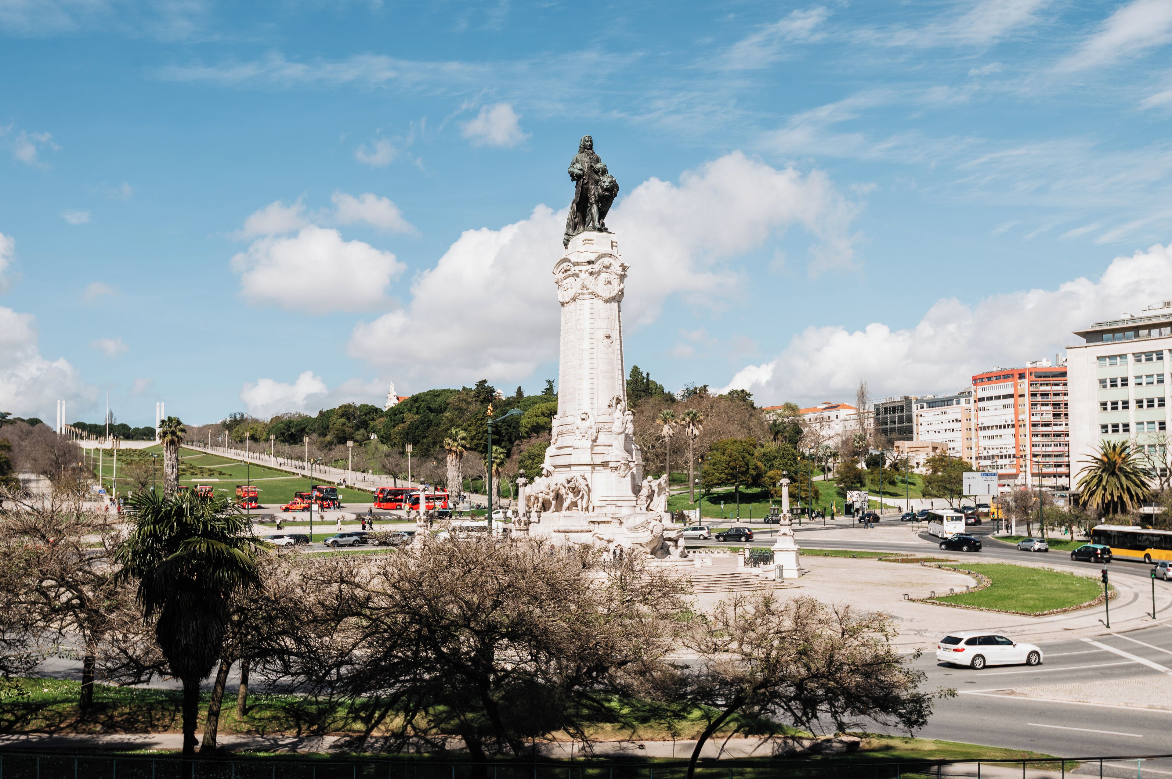 Un monumento alto con una estatua en la cima, rodeado de árboles y una calle concurrida con vehículos.