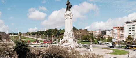 Un monumento alto con una estatua en la cima, rodeado de árboles y una calle concurrida con vehículos.