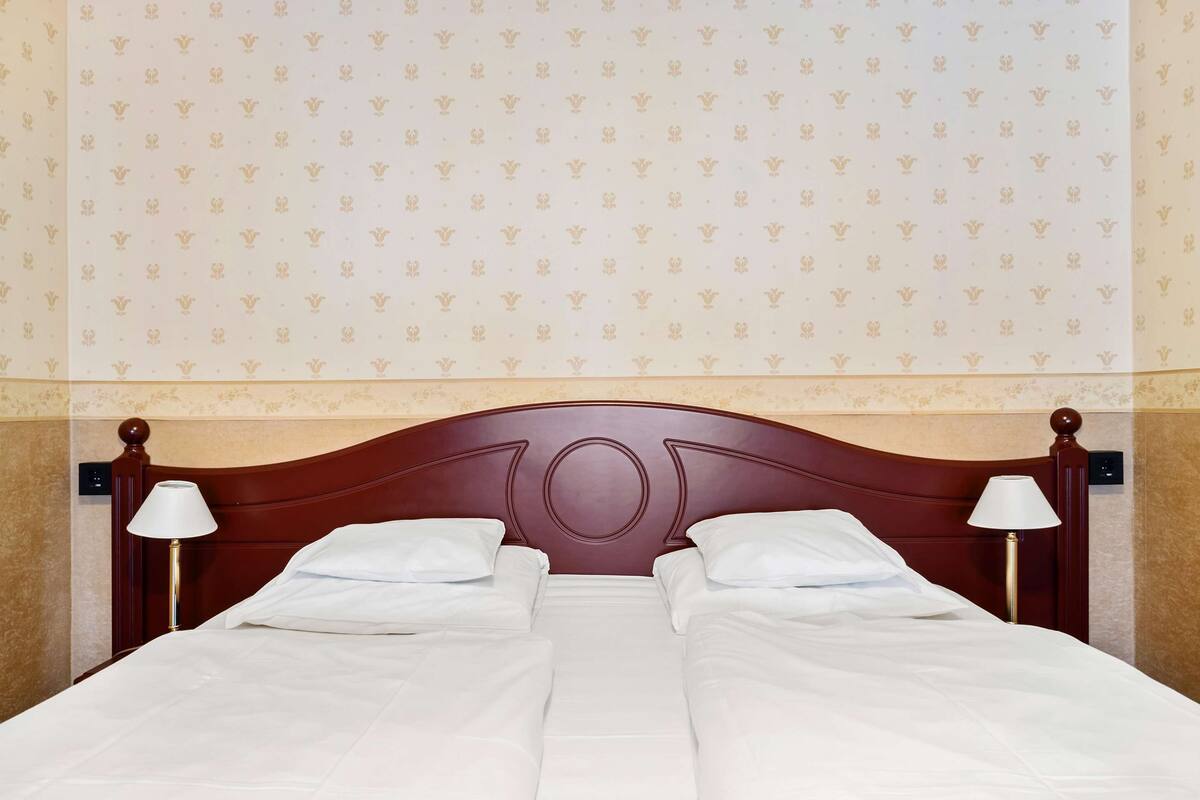 Un lit double avec du linge de lit blanc, une tête de lit en bois, deux lampes de chevet et un mur décoré d’un motif.