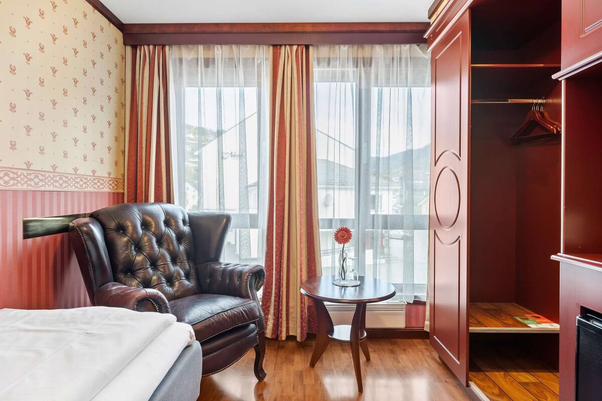 Une chambre d’hôtel avec un fauteuil en cuir, une petite table ronde ornée d’un vase et une armoire avec une tringle pour suspendre les vêtements.