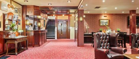 A hotel lobby with a red carpet, wooden furniture, a reception desk, and a staircase.
