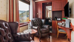 Una habitación con dos sillones de cuero, una mesita redonda pequeña, un escritorio con una silla, un televisor de pantalla plana y vistas a un paisaje montañoso a través de una ventana.