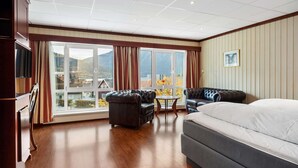 Habitación de hotel con una cama grande, un sofá, una mesita y un televisor. Cuenta con una ventana con cortinas y vistas a edificios y montañas.