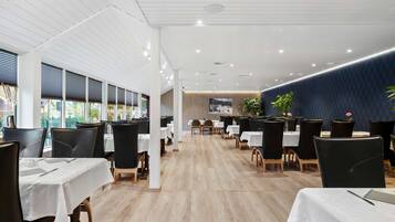 Un restaurante moderno con mesas blancas y sillas negras, suelo de madera y vistas al exterior a través de amplios ventanales.