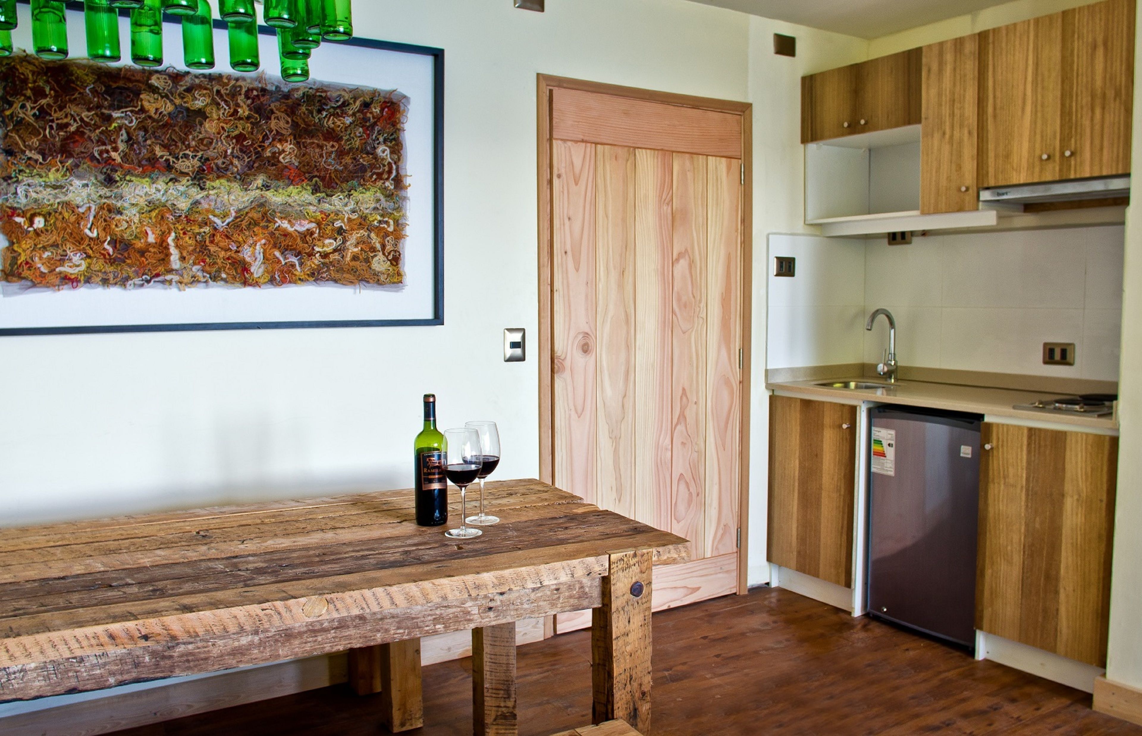 A kitchen with wooden cabinets, a wooden table with a bottle and glass, and a wall art piece.