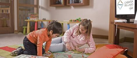 Dos niños jugando con un juego de mesa sobre una alfombra en una habitación con un televisor que muestra el logotipo del Hotel Cabaña del Lago.
