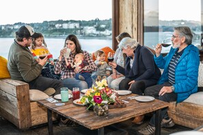 Un groupe de personnes est assis sur un banc en bois près d’une fenêtre donnant sur un plan d’eau. Devant eux, une table est dressée avec des assiettes, des verres et un vase rempli de fleurs.
