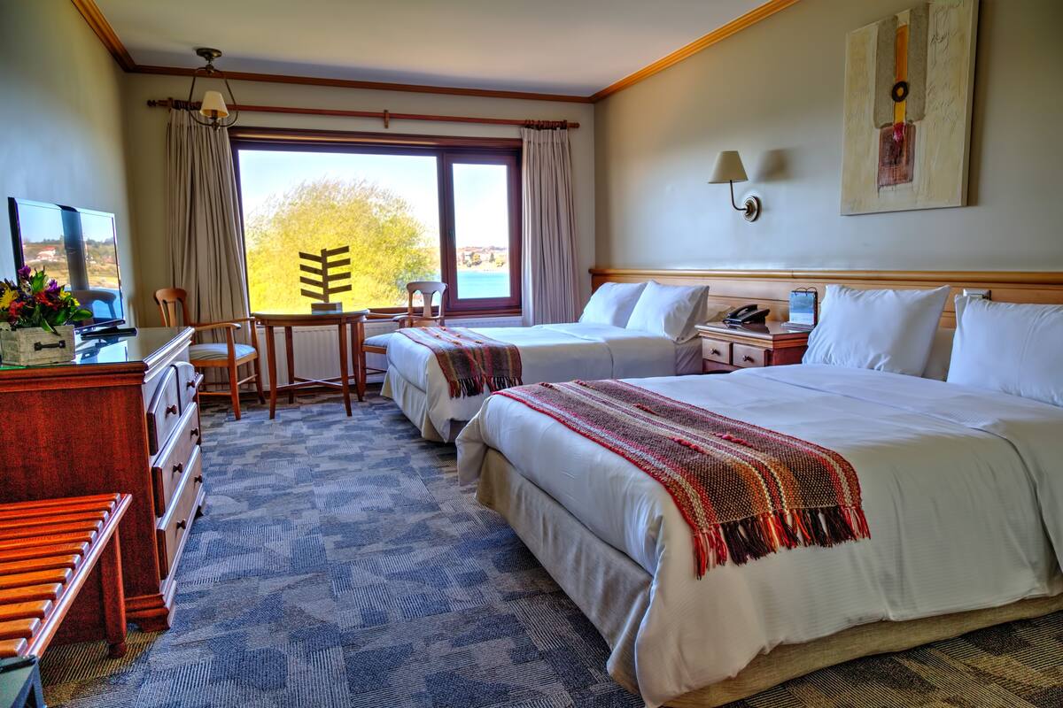 A hotel room with two beds, a desk, a chair, a TV, and a view of the outdoors.