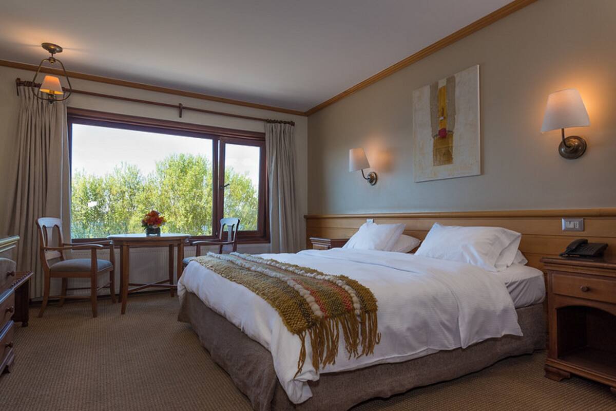 A hotel room with a large bed, a desk with a chair, a window with curtains, and two wall-mounted lamps.