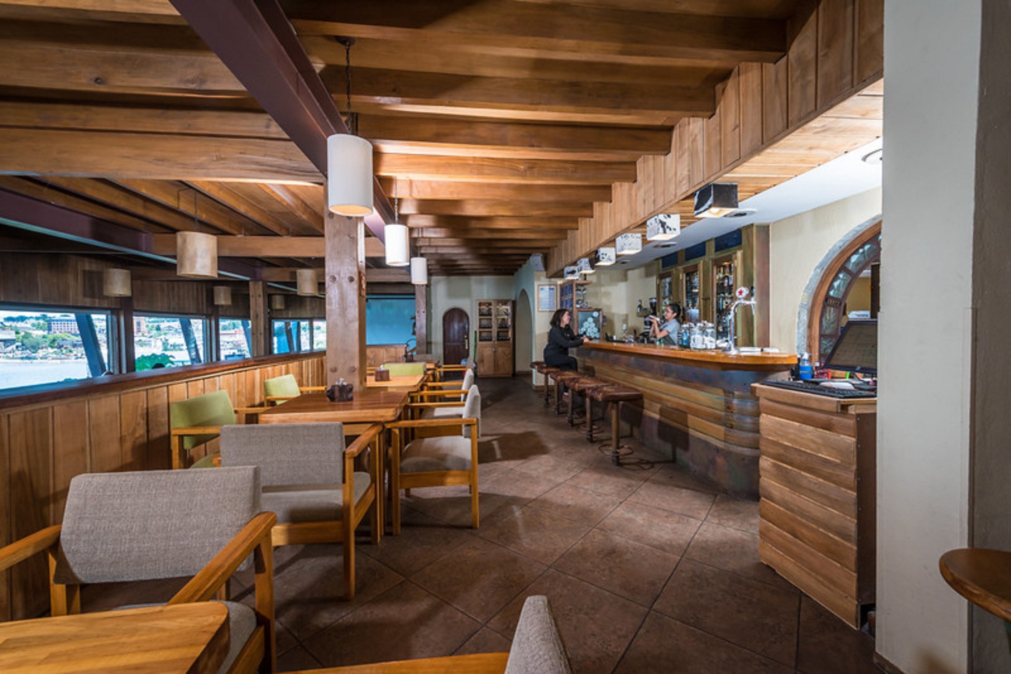 A restaurant with wooden furniture, a bar area, and large windows overlooking a scenic view.