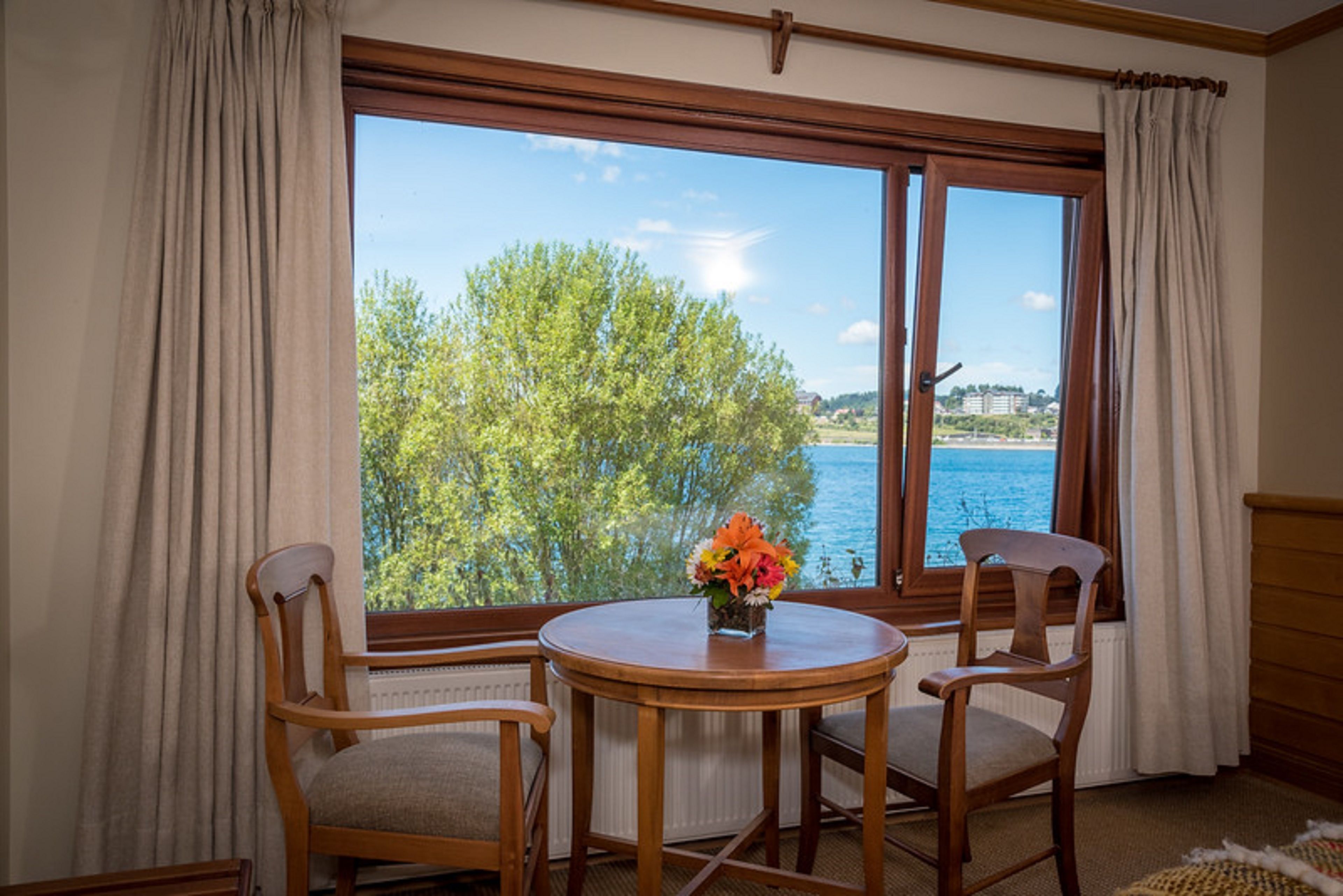 Una habitación con un ventanal amplio, dos sillas de madera, una mesita redonda con un jarrón de flores y vistas a un cuerpo de agua y árboles.