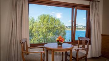 Una habitación con un ventanal amplio, dos sillas de madera, una mesita redonda con un jarrón de flores y vistas a un cuerpo de agua y árboles.