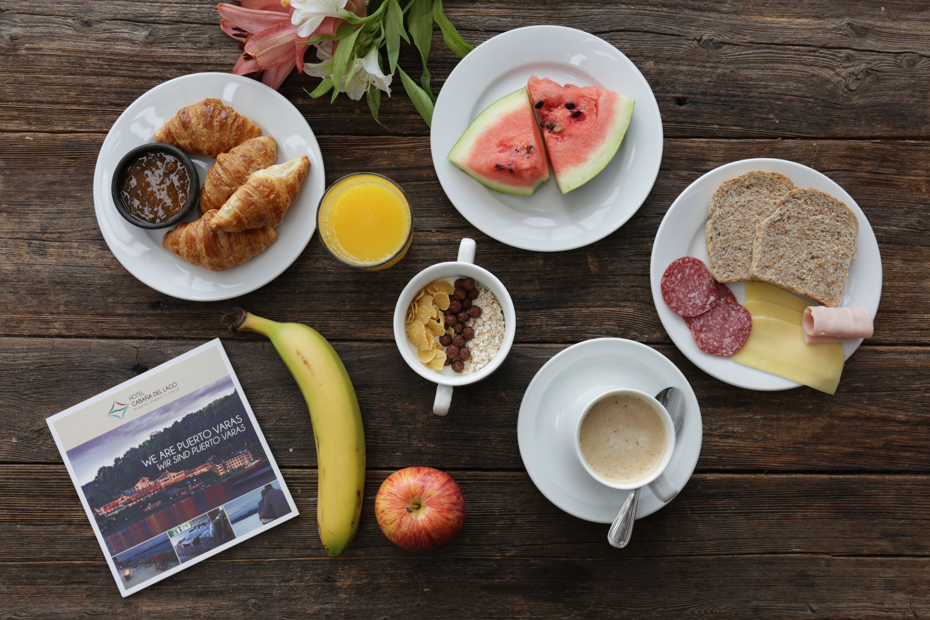 A breakfast setting with croissants, watermelon, bread, cheese, salami, a banana, an apple, cereal, orange juice, coffee, and a brochure on a wooden table.