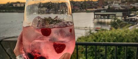 A hand holding a glass with a pink drink, a strawberry, and ice cubes, overlooking a river and cityscape at sunset.