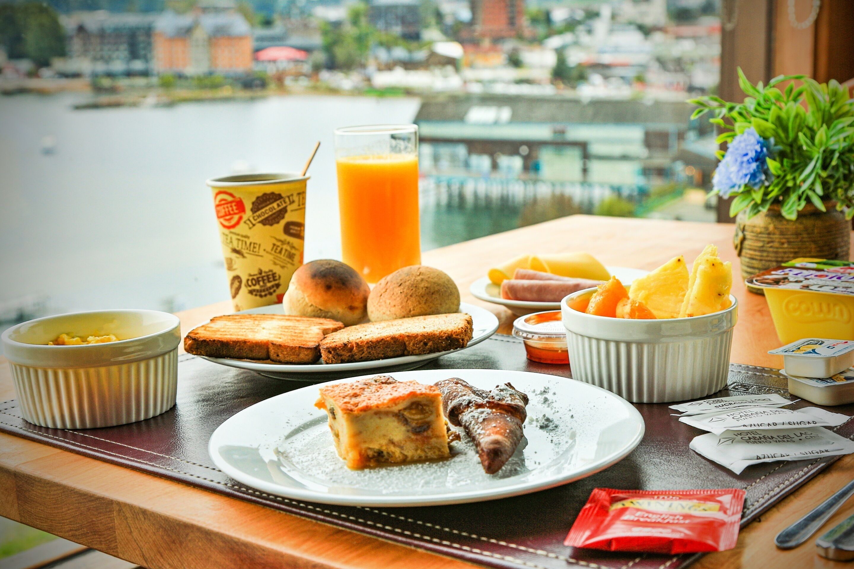 A breakfast table with a variety of food items, including toast, fruit, and a plate with a piece of cake and a sausage. There is a glass of orange juice and a can of coffee. The table is set on a wooden surface with a view of a body of water and buildings in the background.