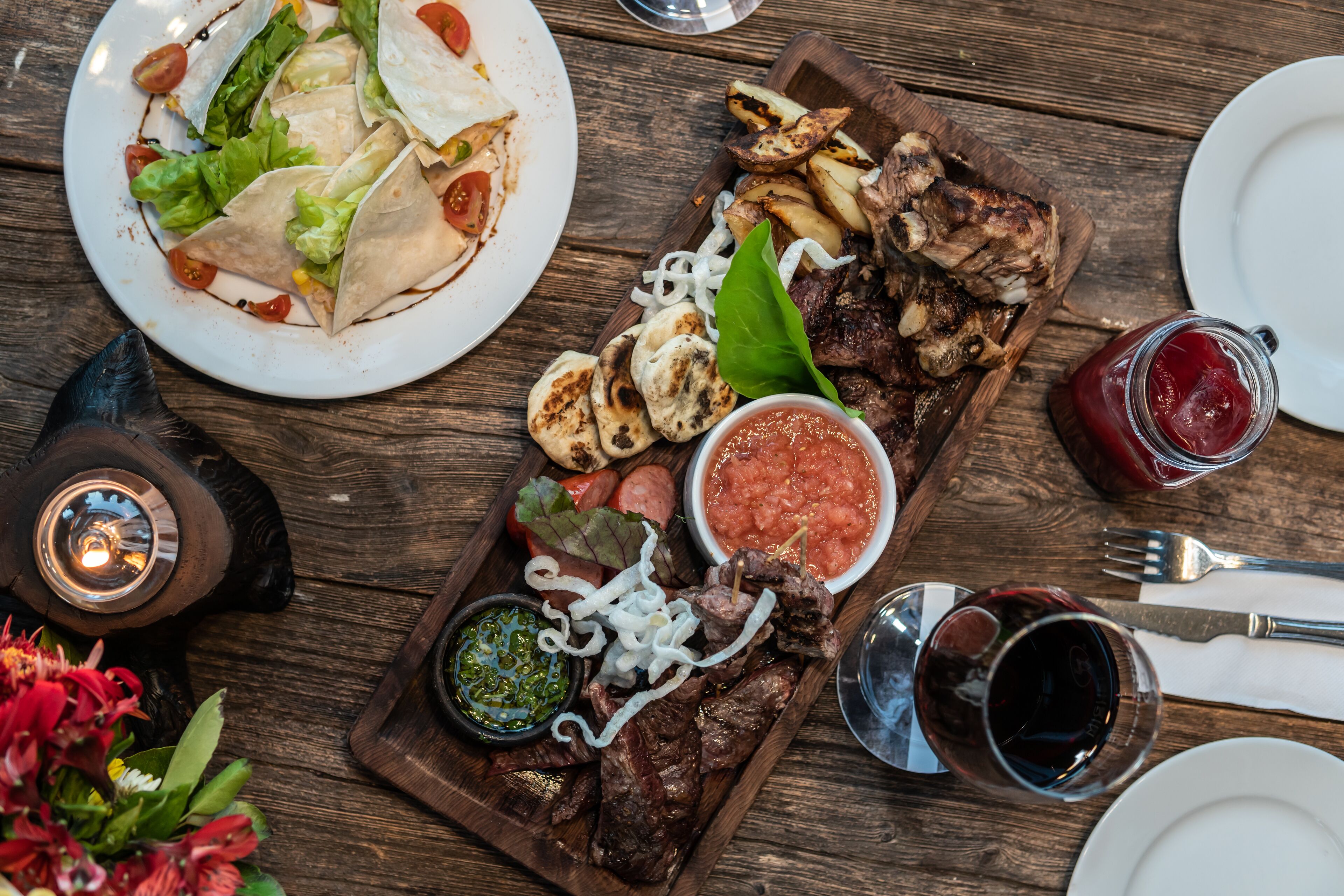 A wooden table with a variety of dishes including tacos, grilled meat, and vegetables, along with condiments and drinks.