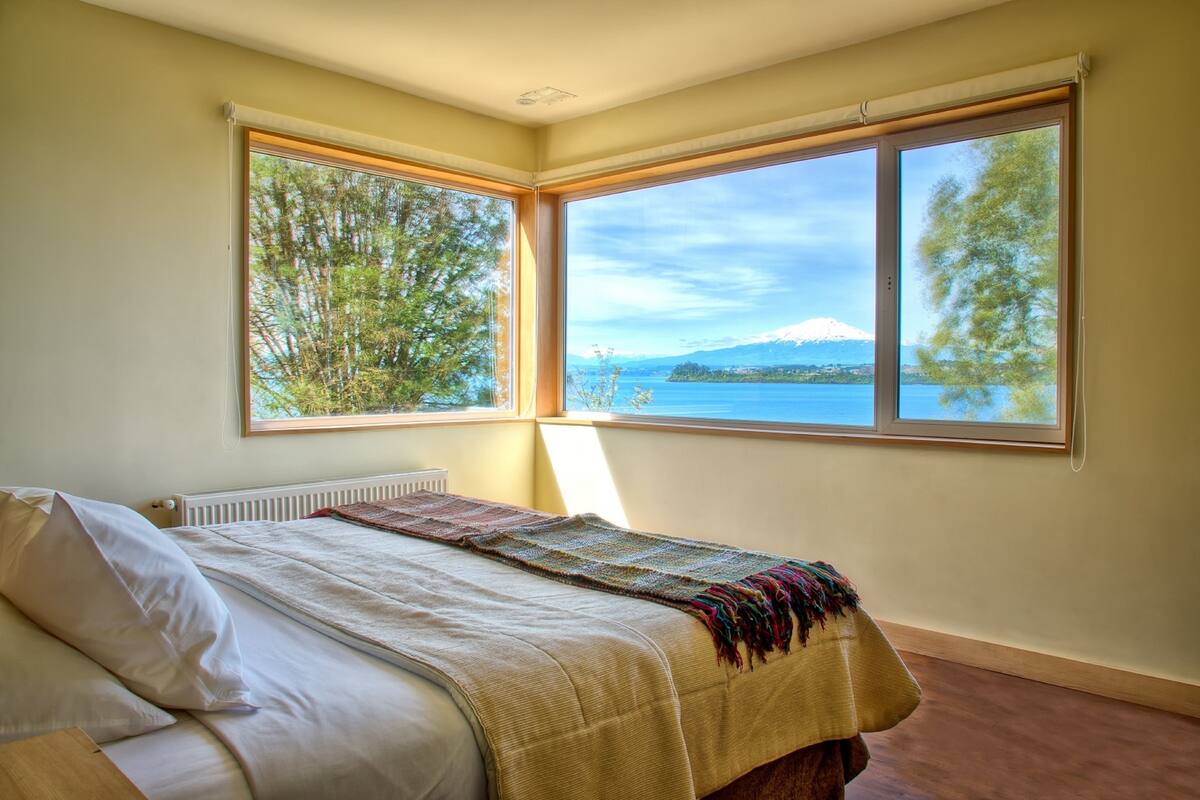 A bedroom with a bed, a radiator, and a large window with a view of a lake and a mountain.