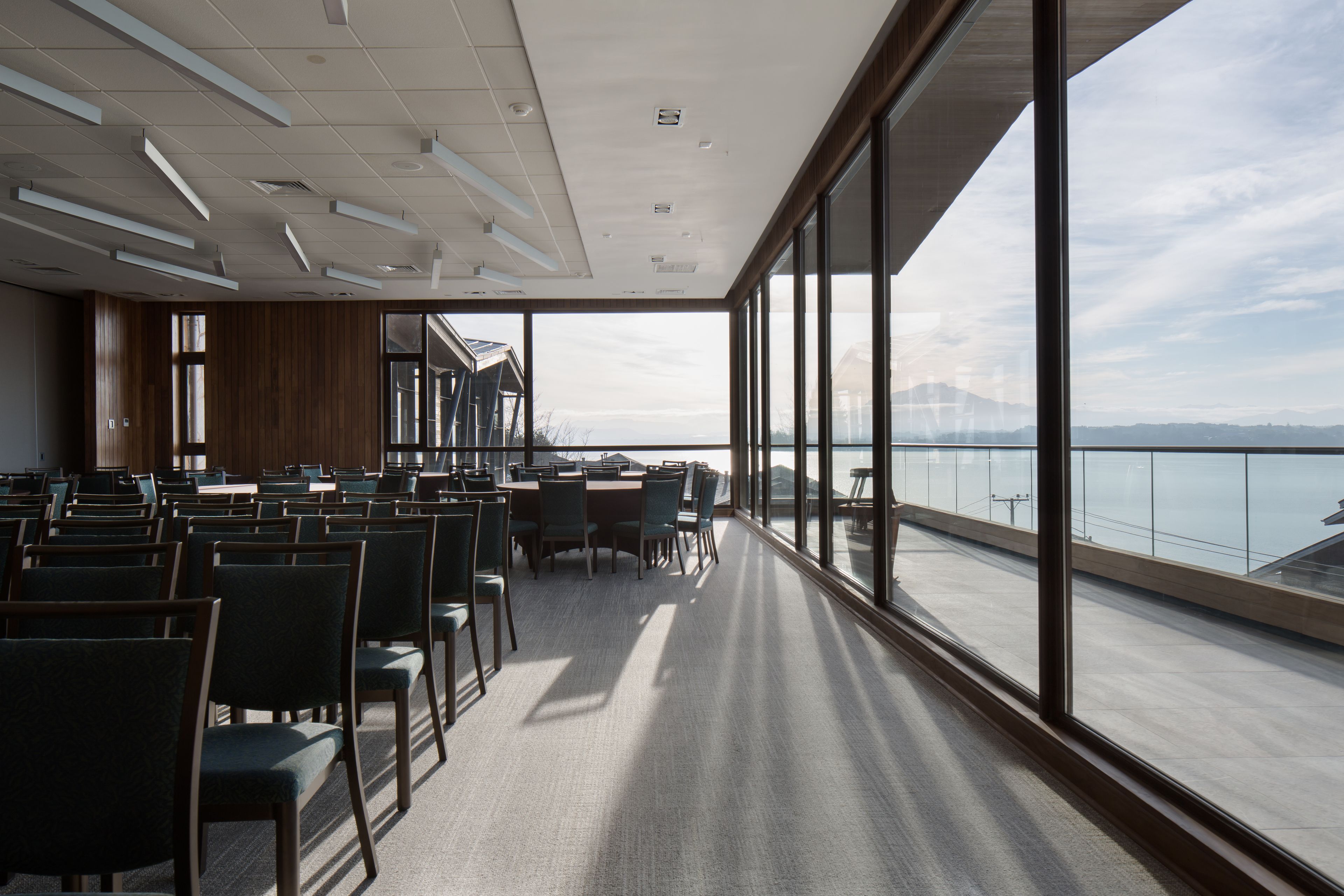 Un salón de conferencias con mesas y sillas, ventanas amplias con vistas al mar y a las montañas, y un techo moderno con iluminación empotrada.