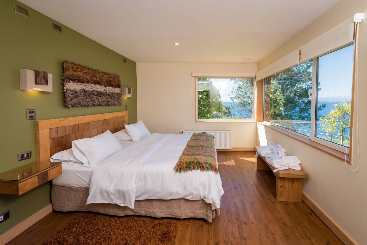 A modern bedroom with a large bed, wooden headboard, a view of the outdoors, and a bench with towels.